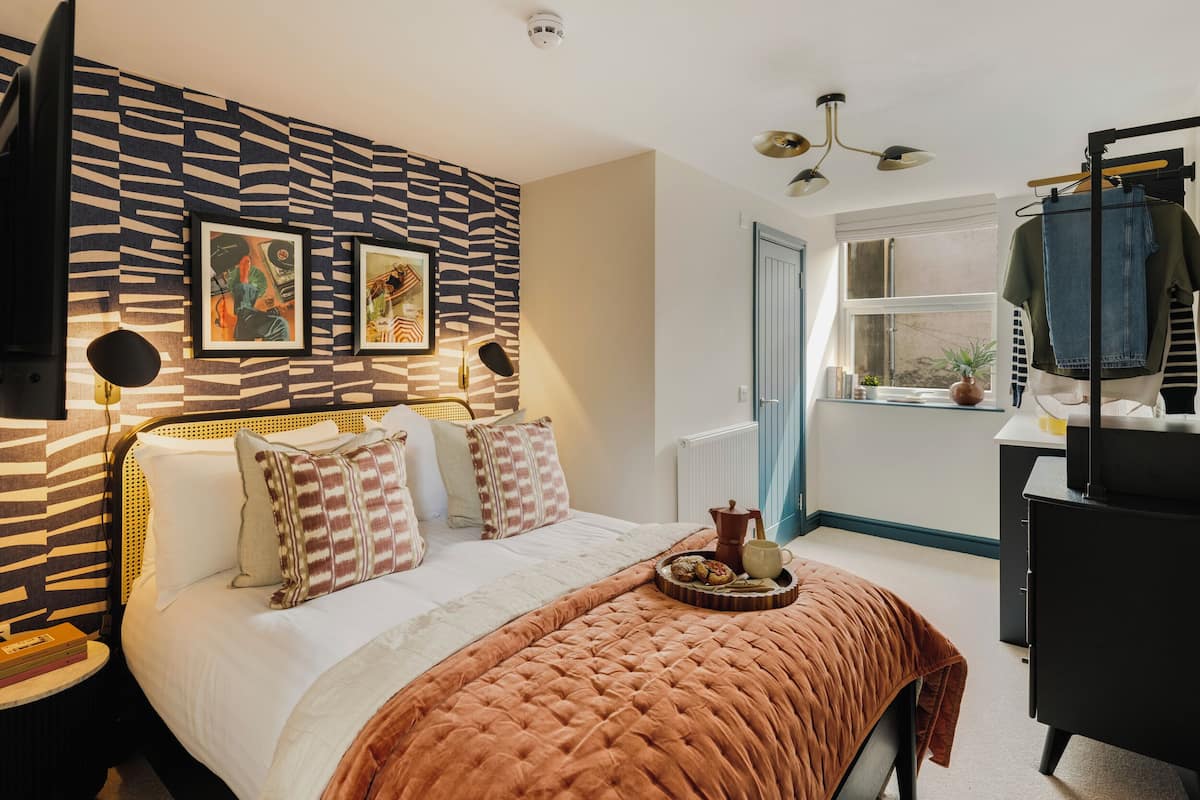 Un dormitorio con un cabecero estampado, una cama con varios almohadones, una mesita de noche con libros y una ventana que da a vistas del exterior.
