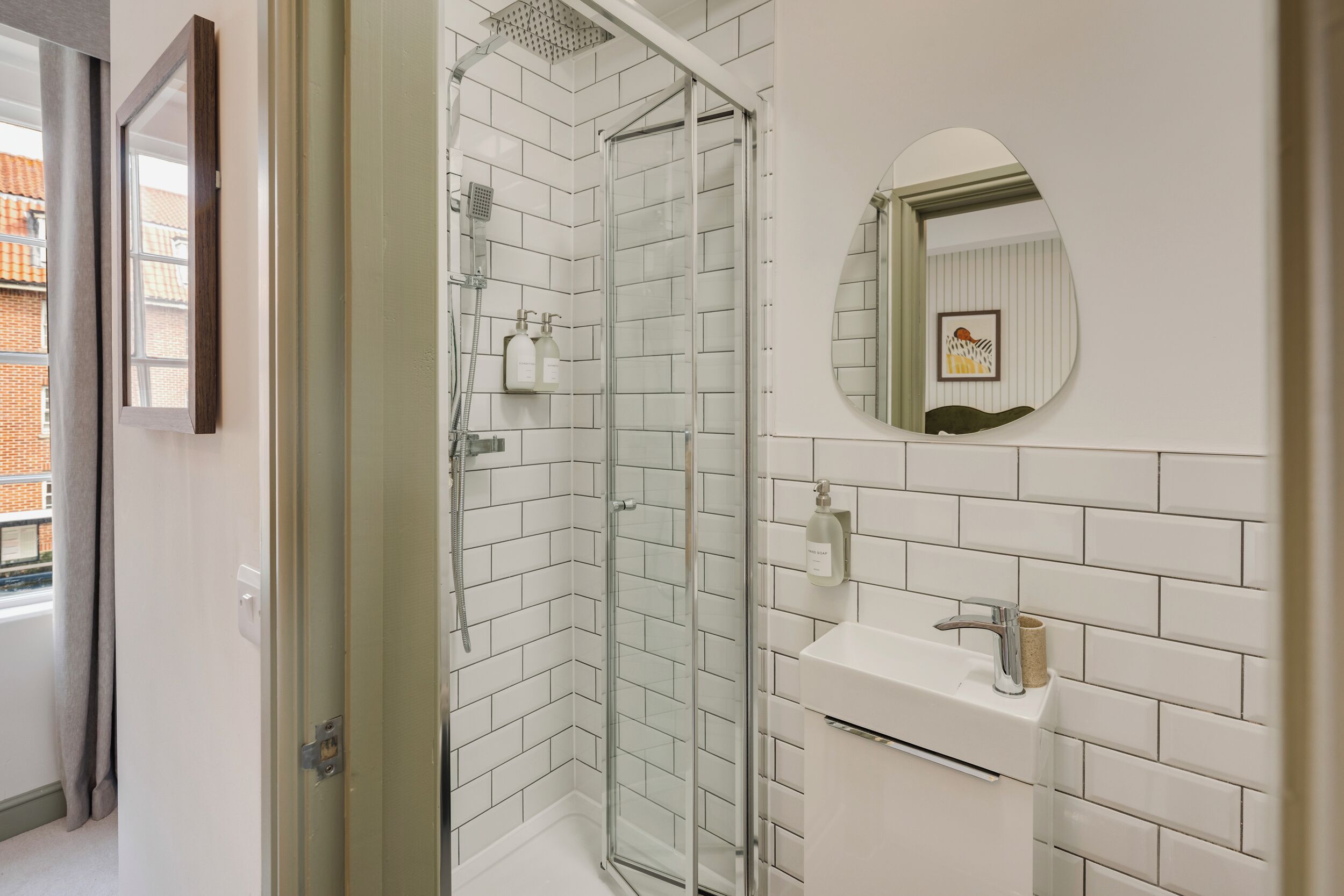 A modern bathroom with white tiles, a glass-enclosed shower, a round mirror, and a white sink.