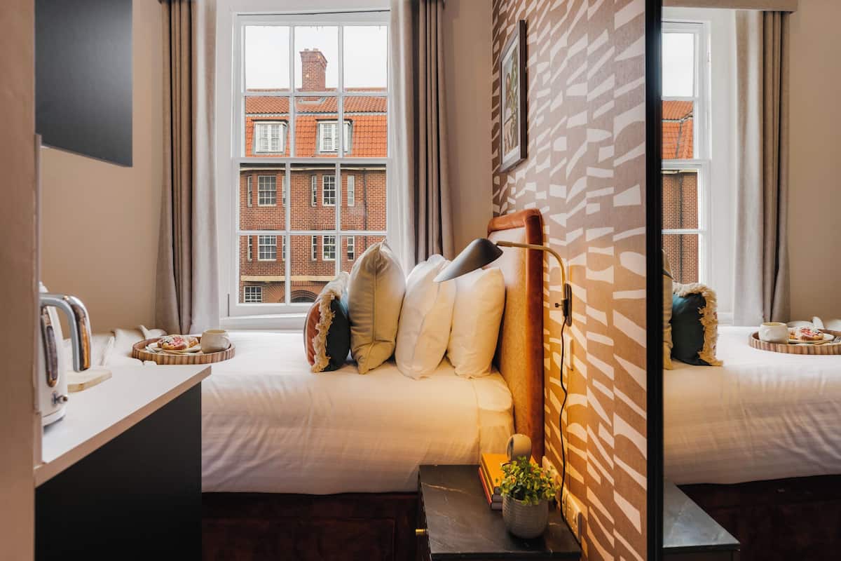 Una habitación de hotel con una cama, una mesita de noche con una bandeja de pasteles, una planta en maceta y vistas a un edificio de ladrillo rojo a través de la ventana.