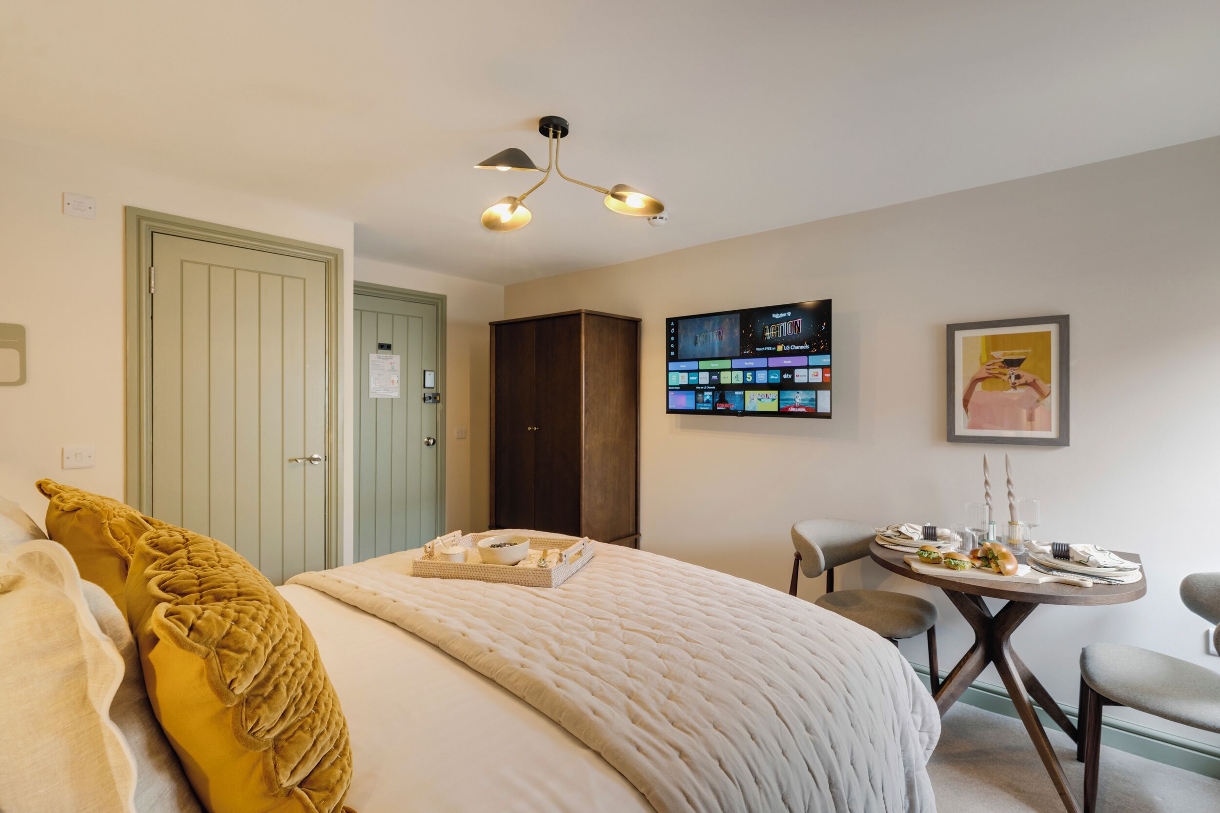 A bedroom with a bed, a bedside table with a tray, a chair, a TV on the wall, and a door.