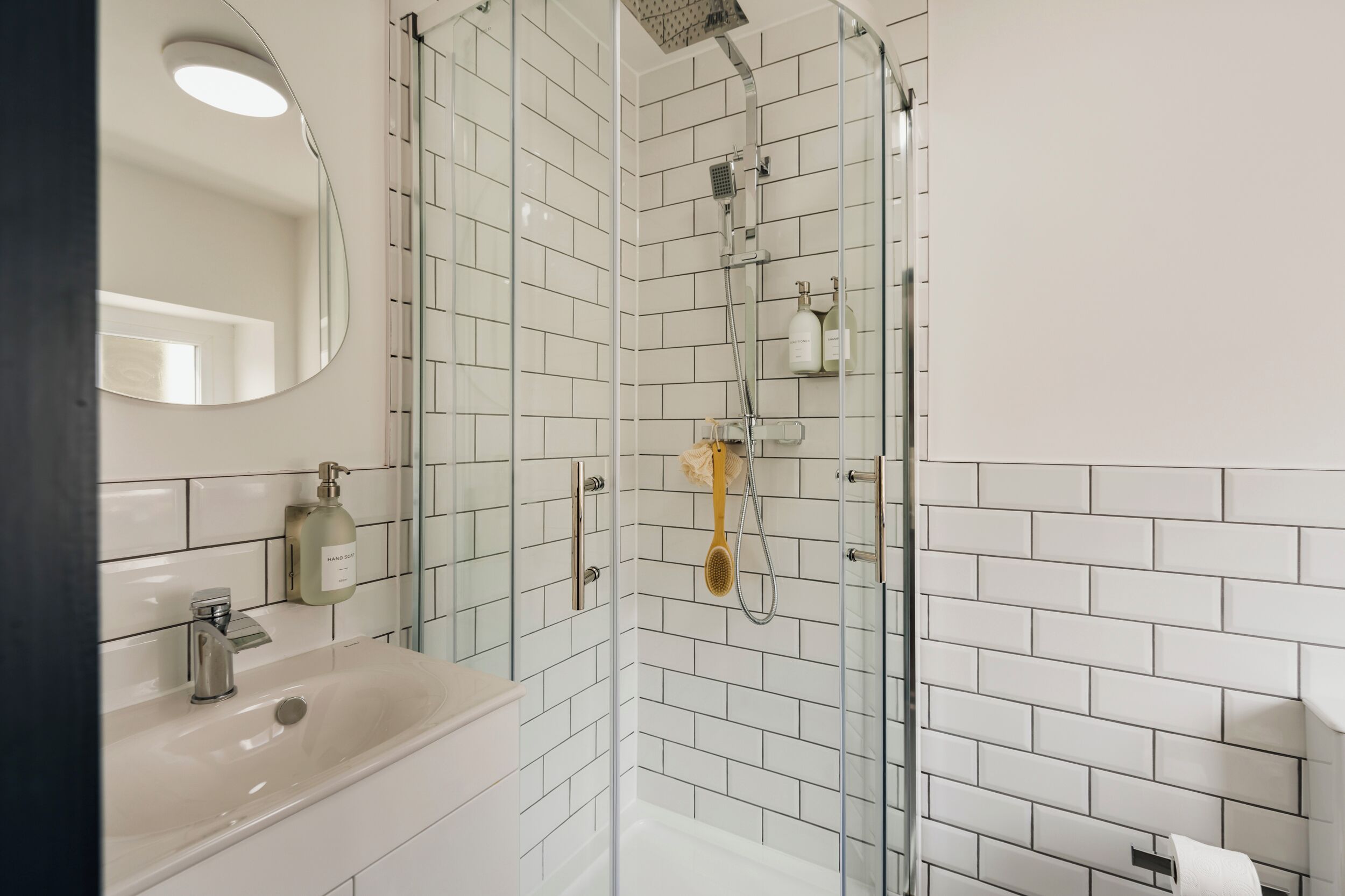 A modern bathroom with a glass-enclosed shower, white tiled walls, a sink with a mirror, and a soap dispenser.