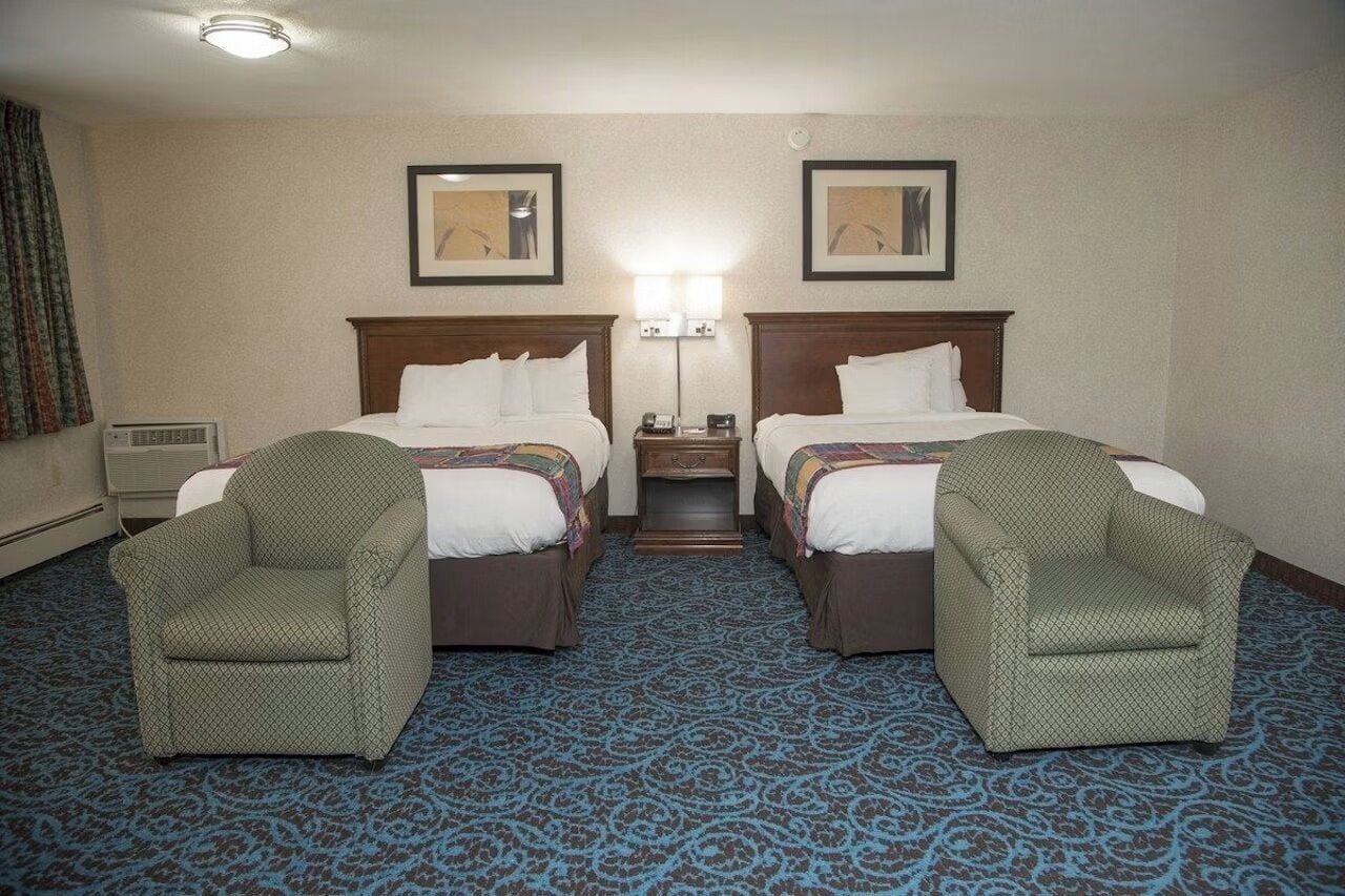 A hotel room with two beds, a nightstand, a lamp, two armchairs, and a carpeted floor.