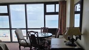 A hotel room with a large window offering a view of the beach and ocean. There is a round table with chairs, a desk with a lamp, and a bed with a white towel.
