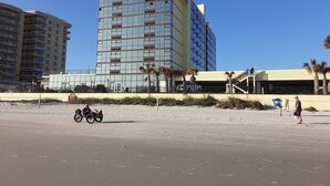 A beachfront hotel with a modern glass building, a sandy beach in the foreground, and palm trees.