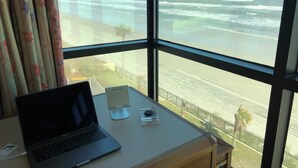A laptop on a desk by a window overlooking a beach with waves.