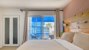 Een hotelkamer met een groot bed, een schuifdeur naar een balkon, een houten hoofdbord en uitzicht op een gebouw en een palmboom.