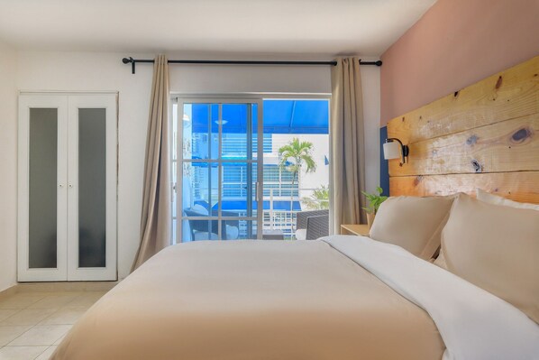 A hotel room with a large bed, a sliding glass door to a balcony, a wooden headboard, and a view of a building and palm tree outside.