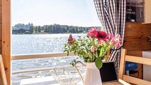A wooden interior with a window overlooking a body of water, a white vase with flowers, and a metal wire basket. - Houseboat BunBo 1060 - Do (Königs Wusterhausen OT Zernsdorf)