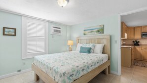 A bedroom with a wooden bed, a nightstand, a lamp, a window with blinds, a painting on the wall, and a view into the kitchen.