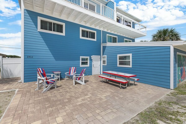 A two-story building with a blue exterior, a balcony, and a patio area with red and white Adirondack chairs and a picnic table.