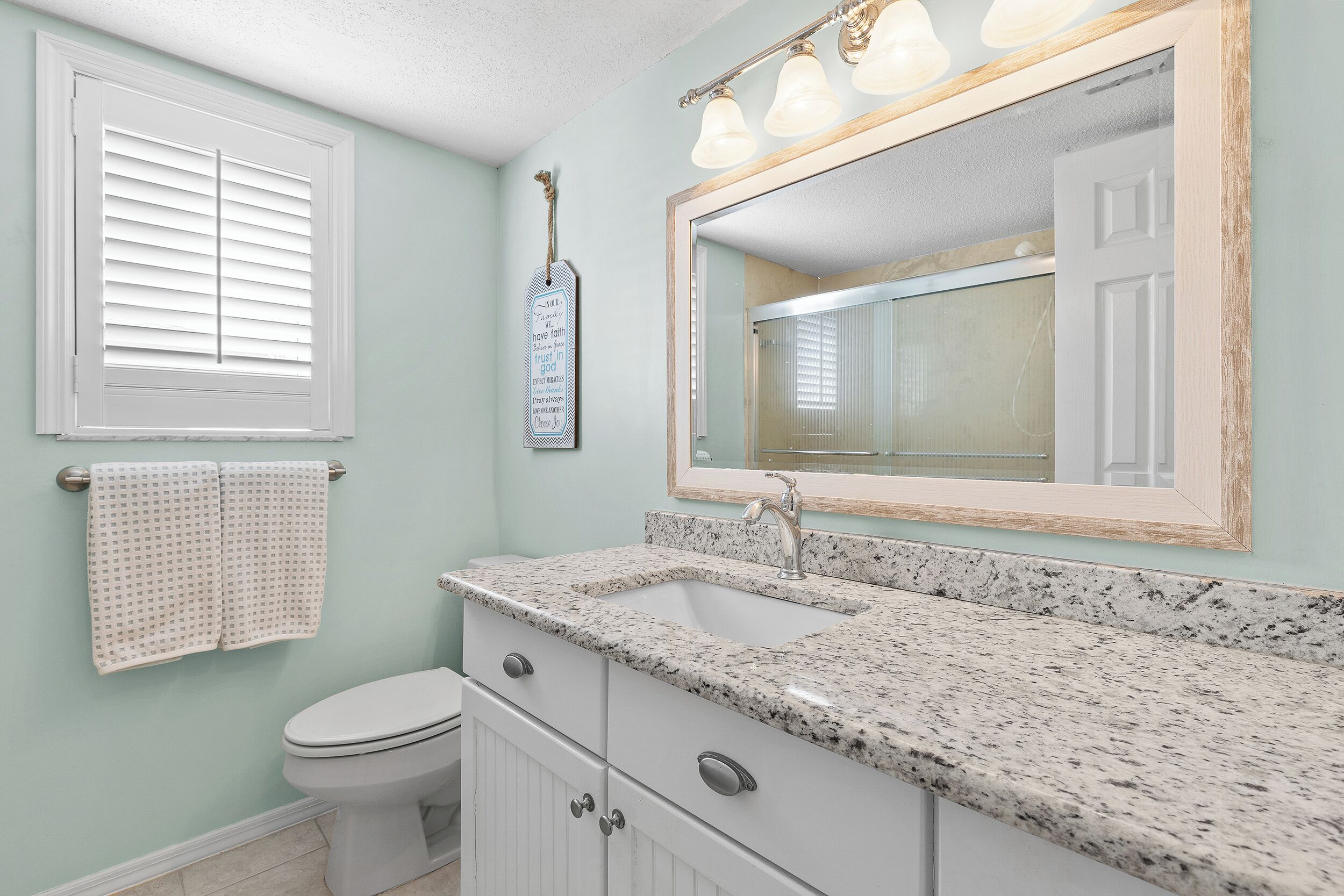 A bathroom with a white toilet, a sink with a granite countertop, a mirror with a wooden frame, and a window with blinds.