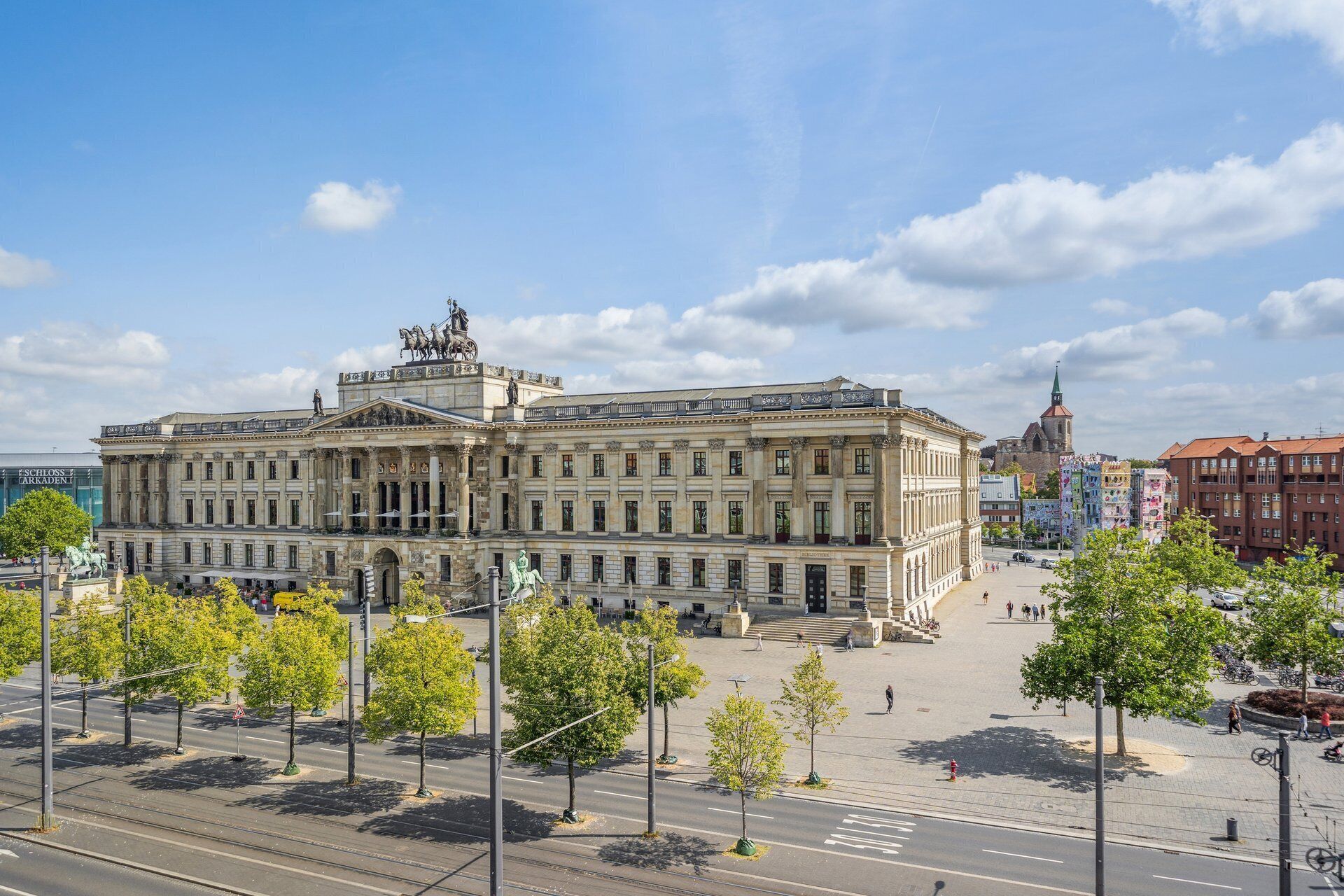 A large, historic building with classical architecture, a prominent statue on the roof, and a surrounding open square with trees and a tram line.