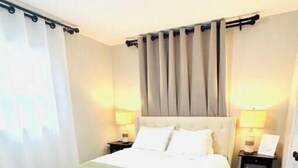 Una habitación de hotel con cama, dos mesitas de noche con lámparas, una ventana con cortinas y suelo de madera.