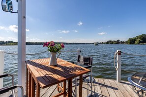 Eine Holzterrasse mit Tisch und Stuhl, einem Blumentopf und Blick auf einen See mit einer Brücke in der Ferne.