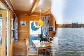 Une cabane avec un intérieur en bois, une table avec des chaises, une lampe suspendue et une vue sur l’eau.