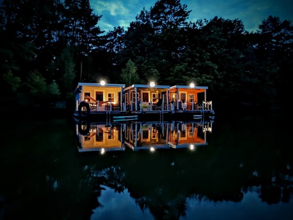 Une rangée de maisons flottantes, reflétées dans l’eau, reliées par une jetée et entourées d’arbres.