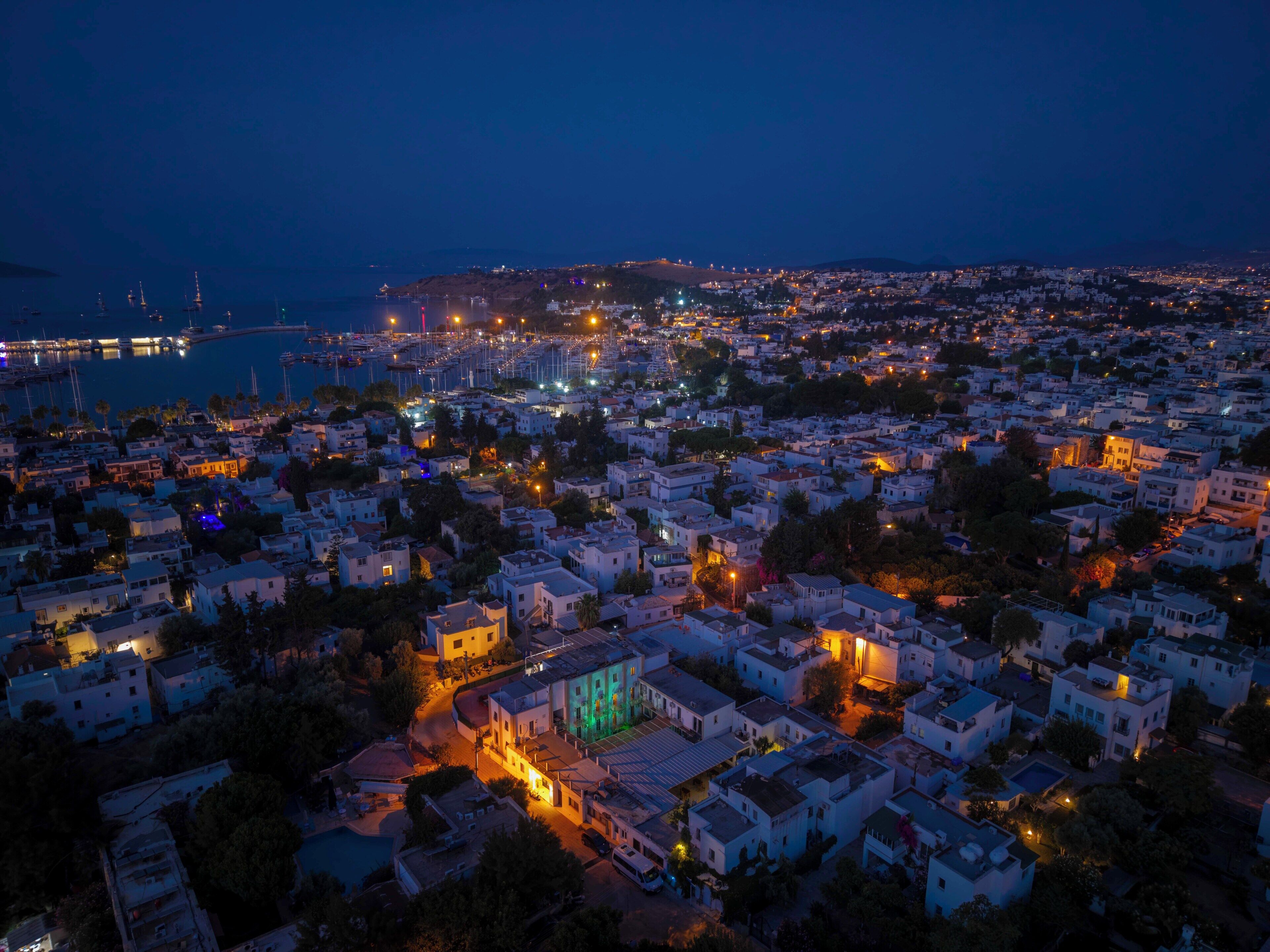 Une ville côtière au crépuscule, avec un port de plaisance, des bâtiments blancs et un immeuble imposant illuminé de lumière verte.