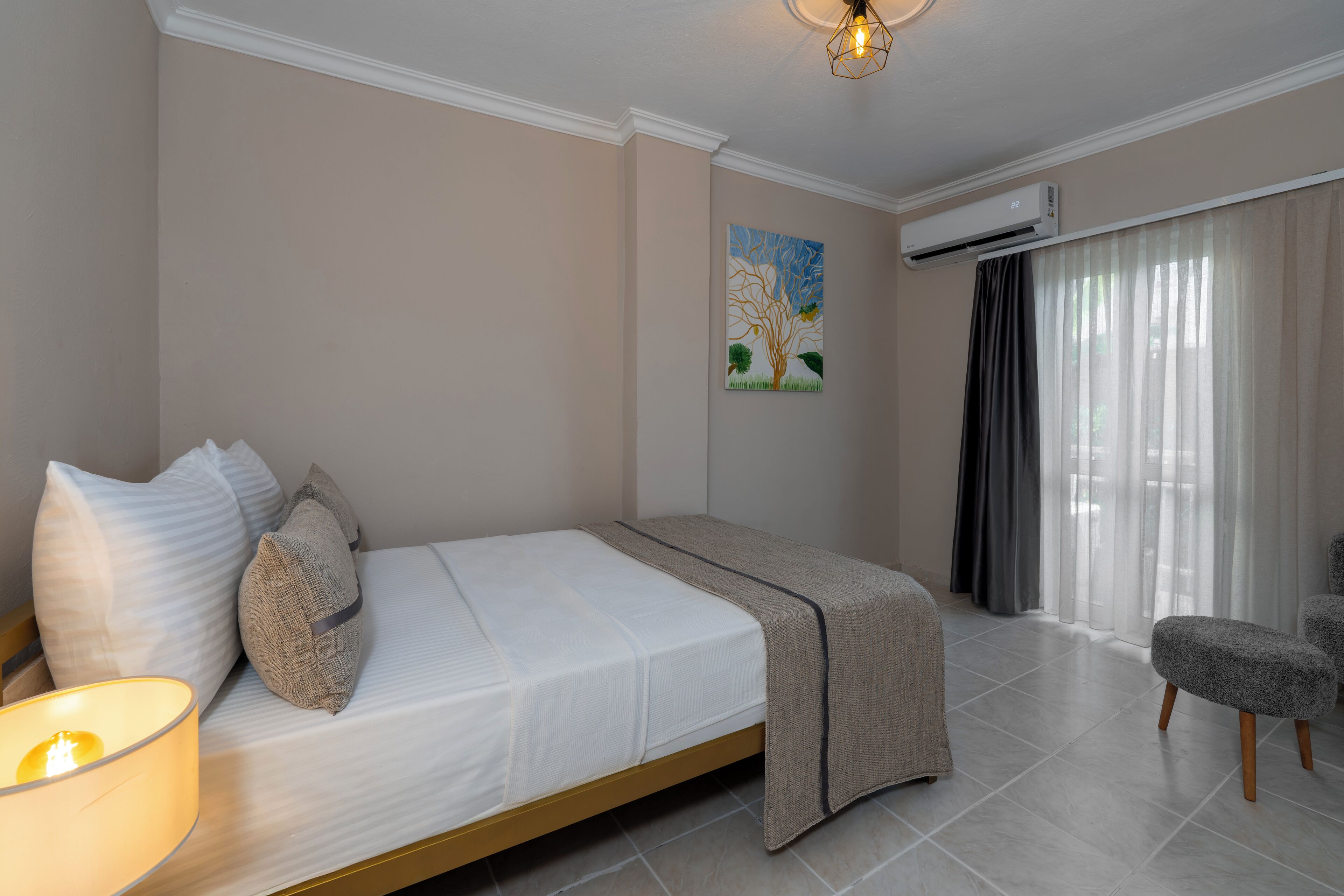 A neatly arranged bedroom with a bed, pillows, a bedside lamp, a chair, and an air conditioning unit.