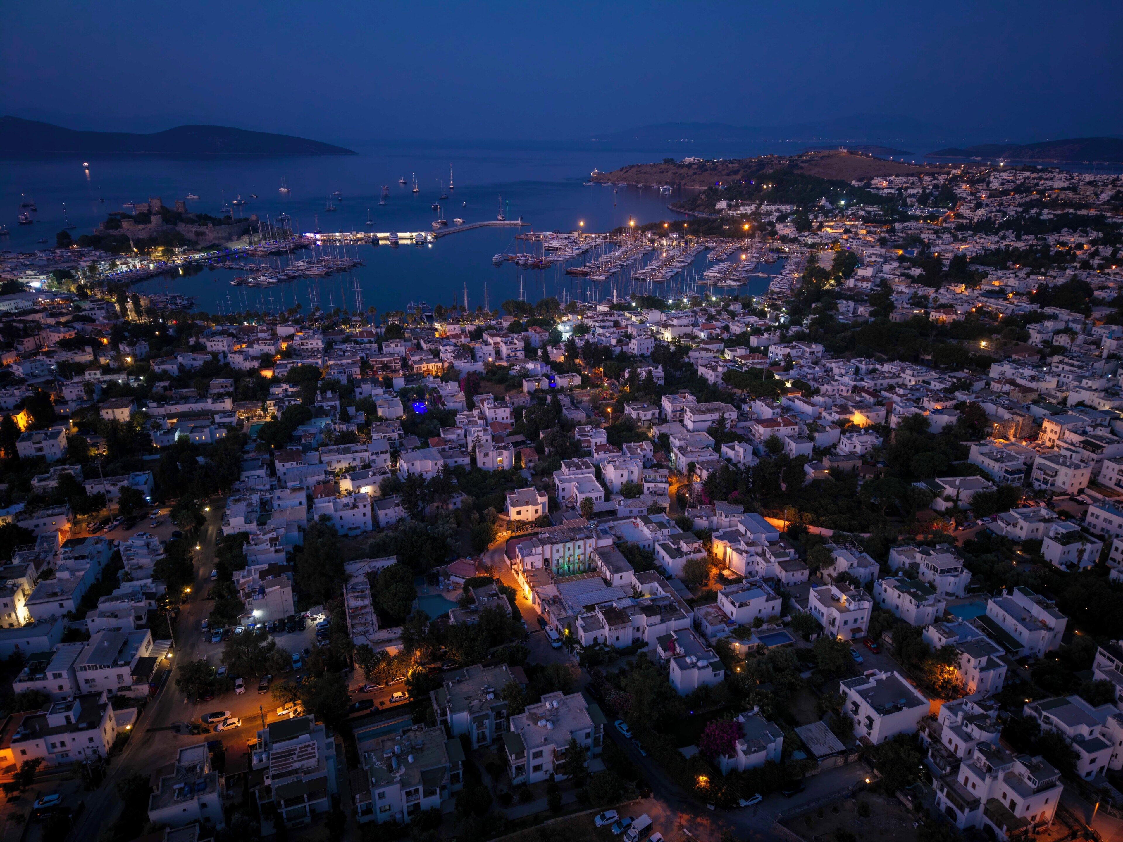 A coastal town at dusk with a marina, residential buildings, and a prominent castle-like structure.