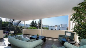 Un espacio exterior moderno para disfrutar al aire libre, con sofá, sillas y una mesa de centro. Una escalera conduce a un nivel superior, y se aprecia una vista de edificios y vegetación al fondo.