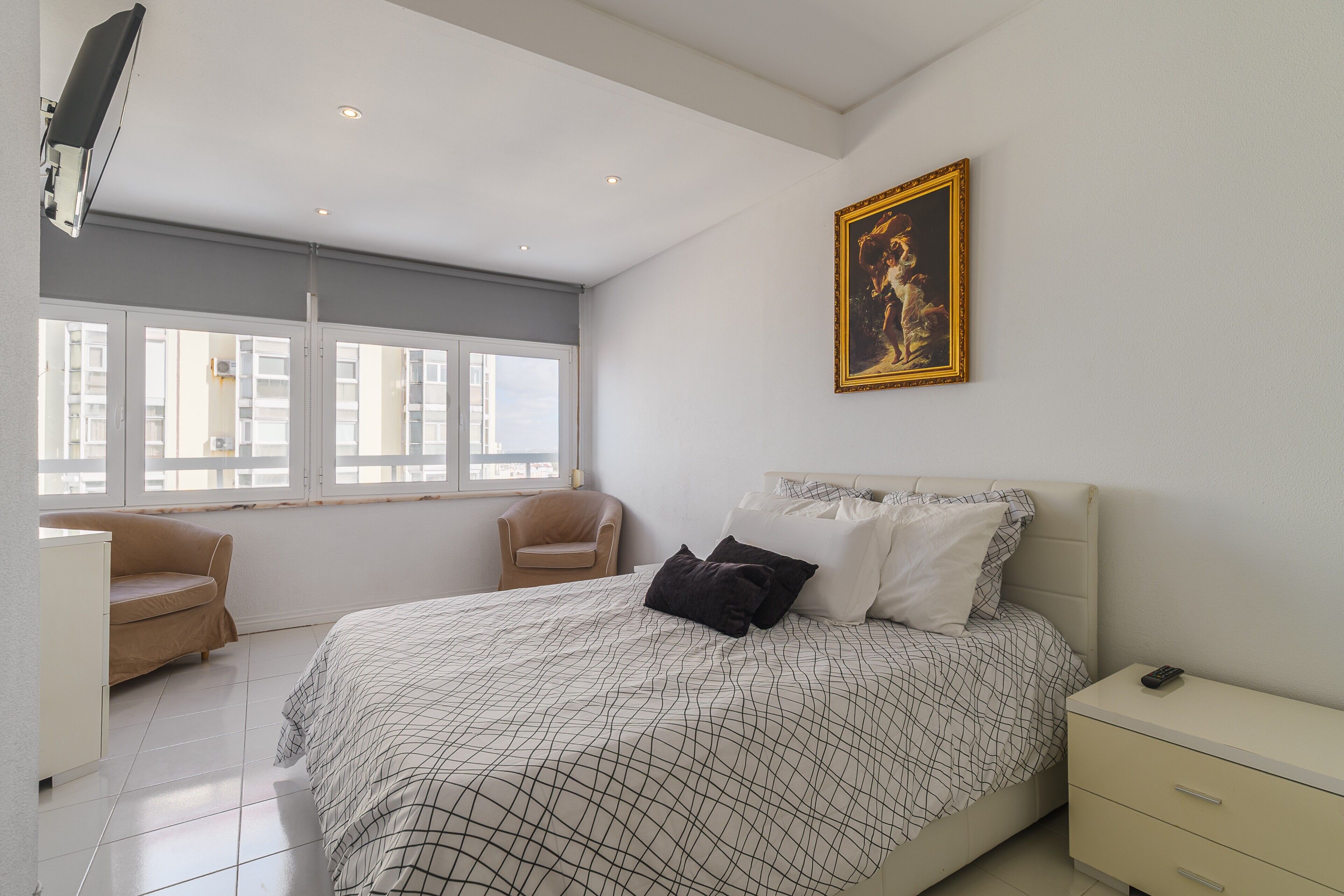 Un dormitorio con una cama, una silla, una mesita de noche, un cuadro y una ventana.