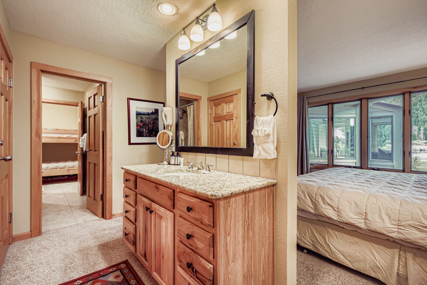 A bedroom with a large window, a wooden vanity with a mirror, and a bunk bed in the adjacent room.