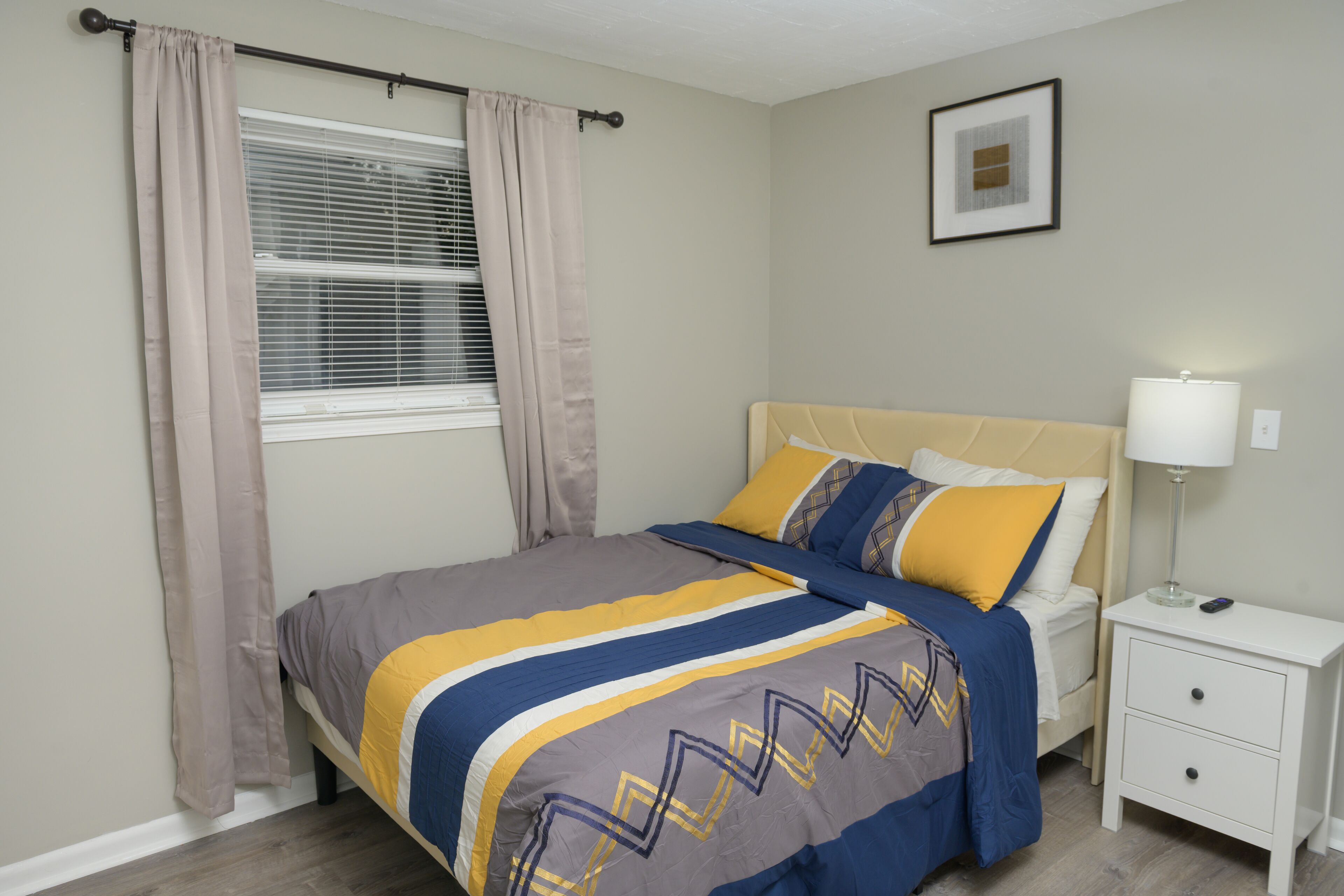 Ein ordentlich bezogenes Bett mit einem gemusterten Überwurf, ein Nachttisch mit Lampe und ein Fenster mit Vorhängen.