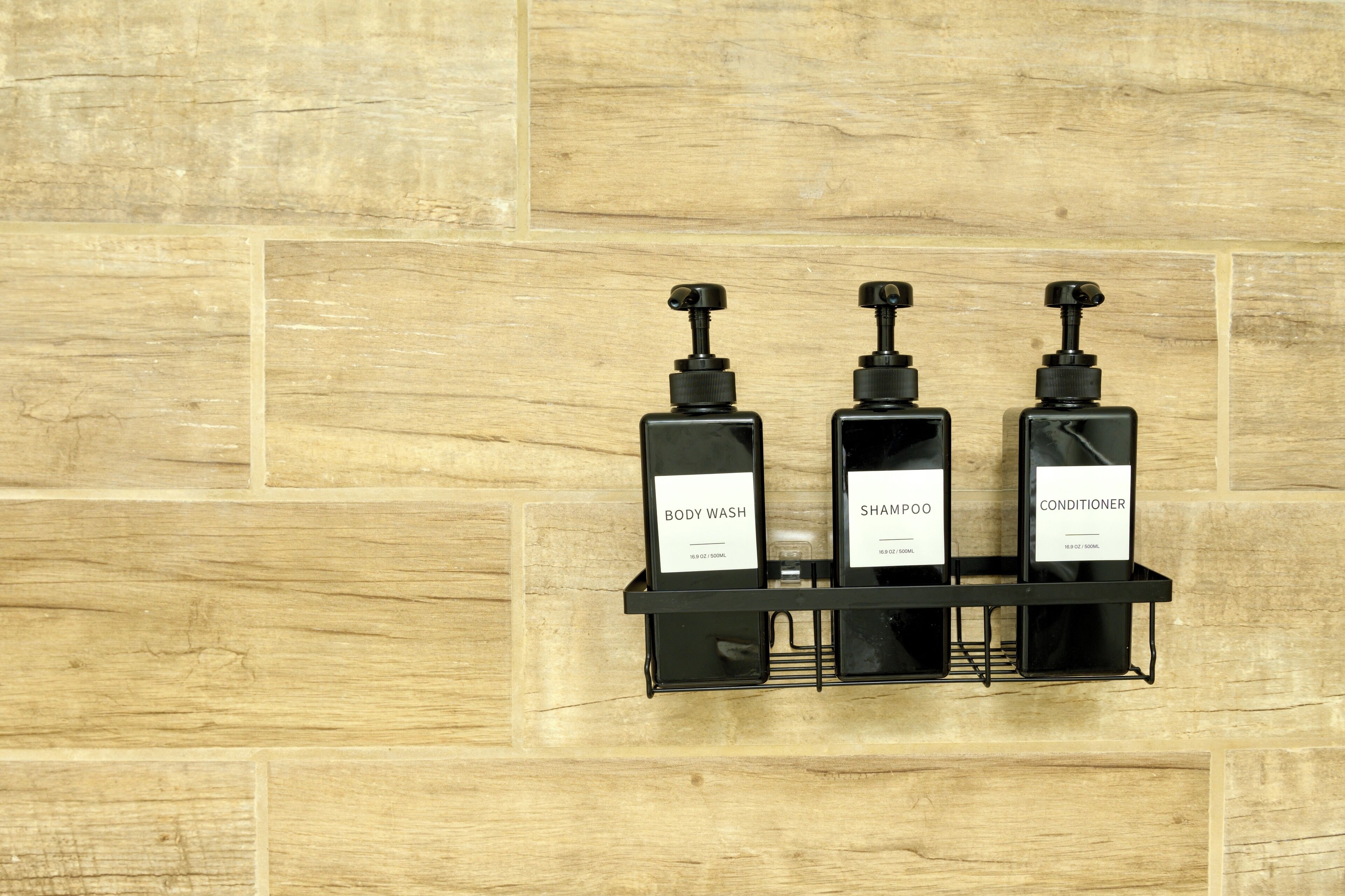 Three black bottles of body wash, shampoo, and conditioner on a black shelf against a wooden wall.