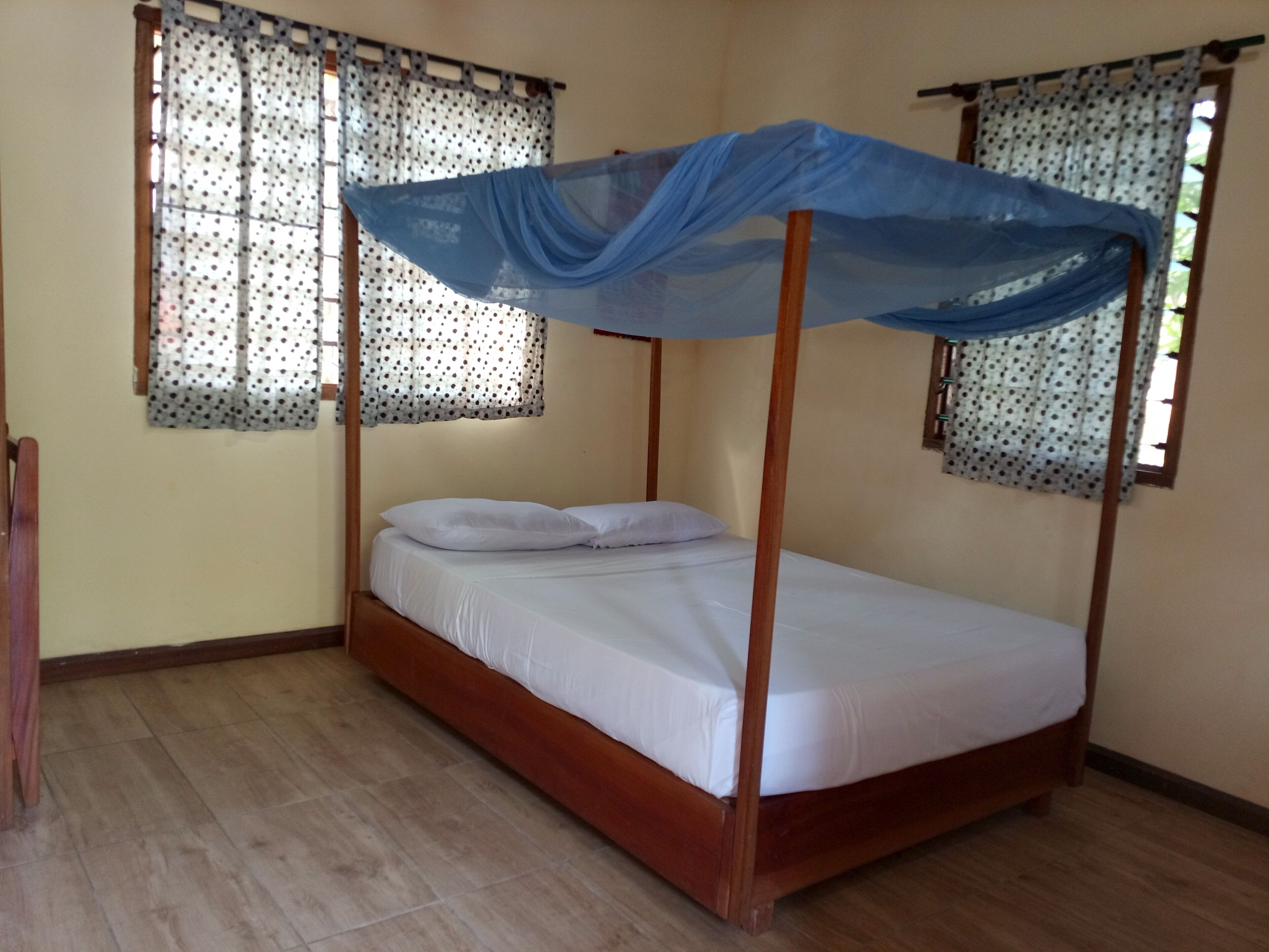 A single wooden bed with a canopy, two windows with patterned curtains, and a wooden floor.