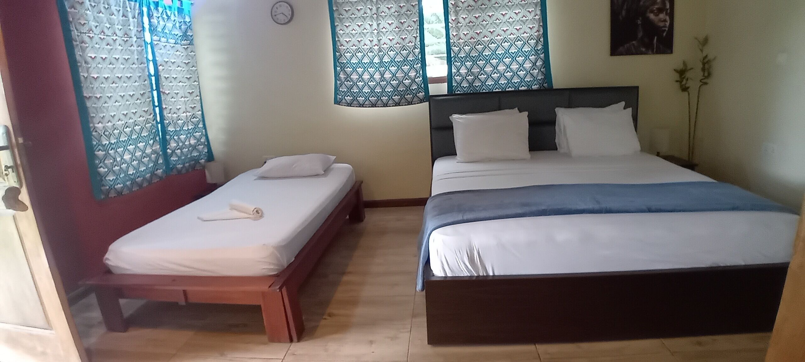 A bedroom with two beds, one larger and one smaller, both with white bedding. There is a clock on the wall and a plant on the right side.