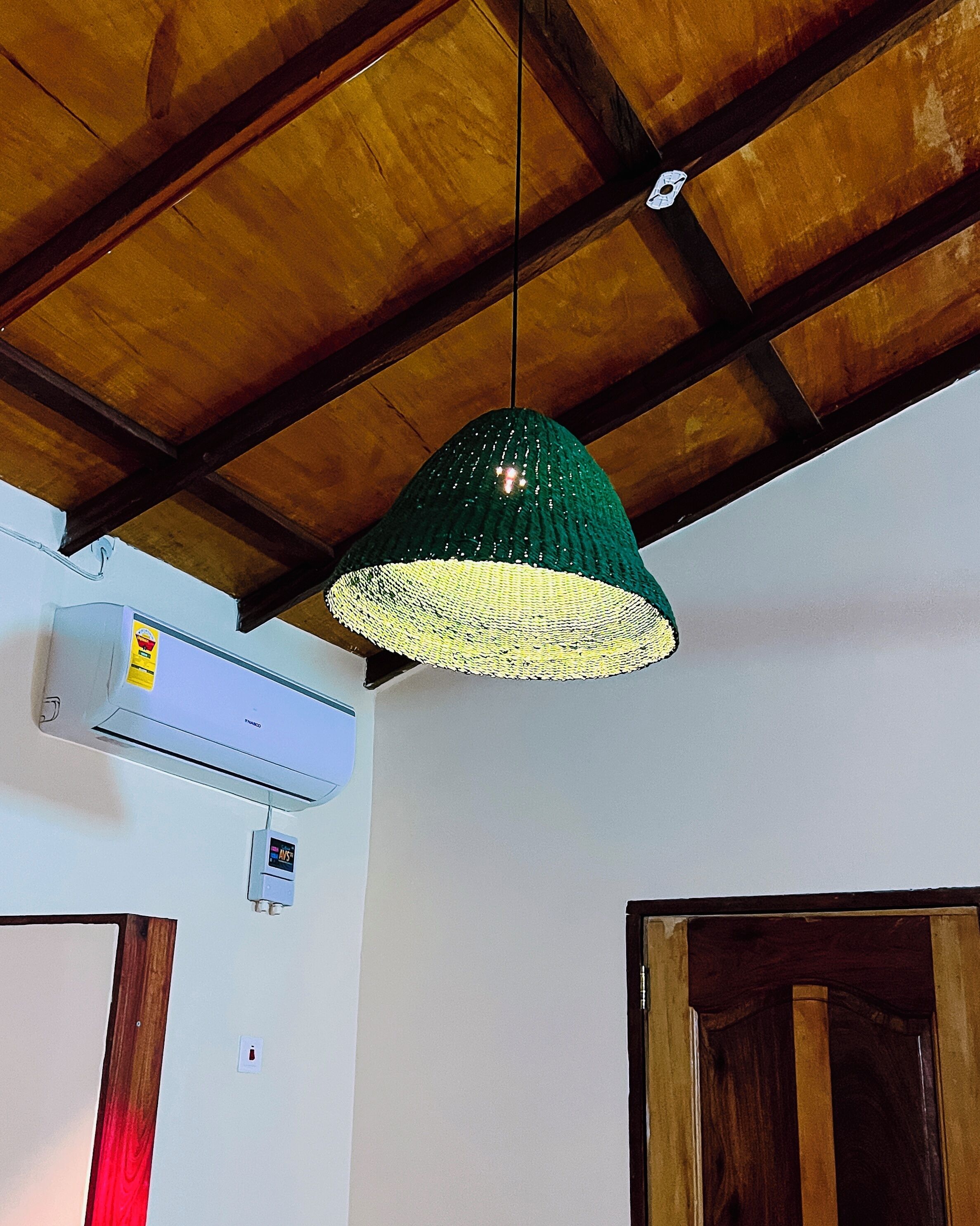A room with a wooden ceiling, a green knitted lamp, an air conditioning unit, and a wooden door.