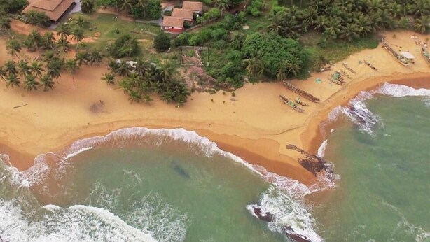 An aerial view of a coastal area with a sandy beach, boats, and a small settlement with houses and palm trees. - Ko-Sa Beach Resort (Ampenyi)