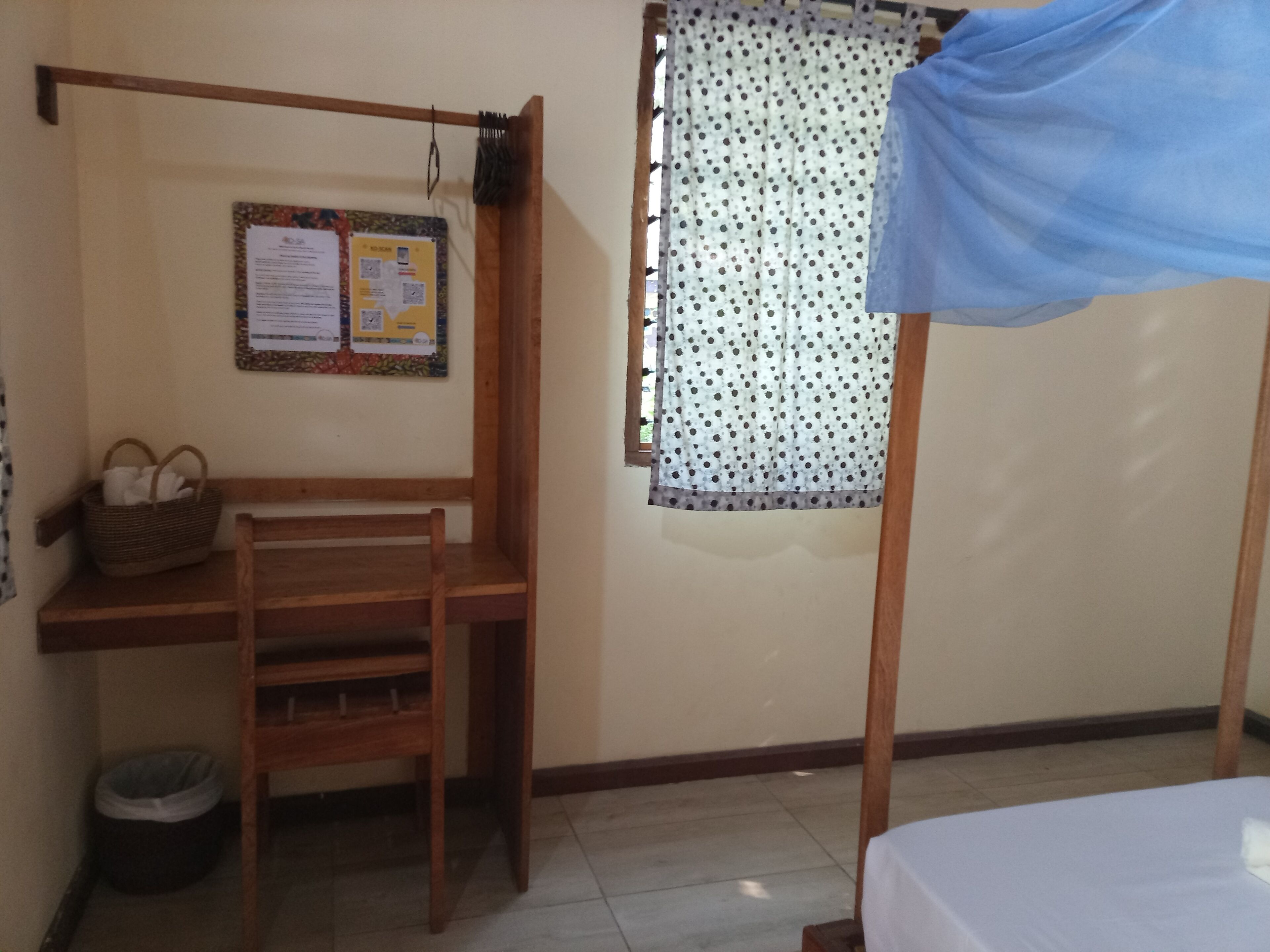 A small room with a wooden desk, a chair, a bed, a window with a curtain, and a bulletin board on the wall.