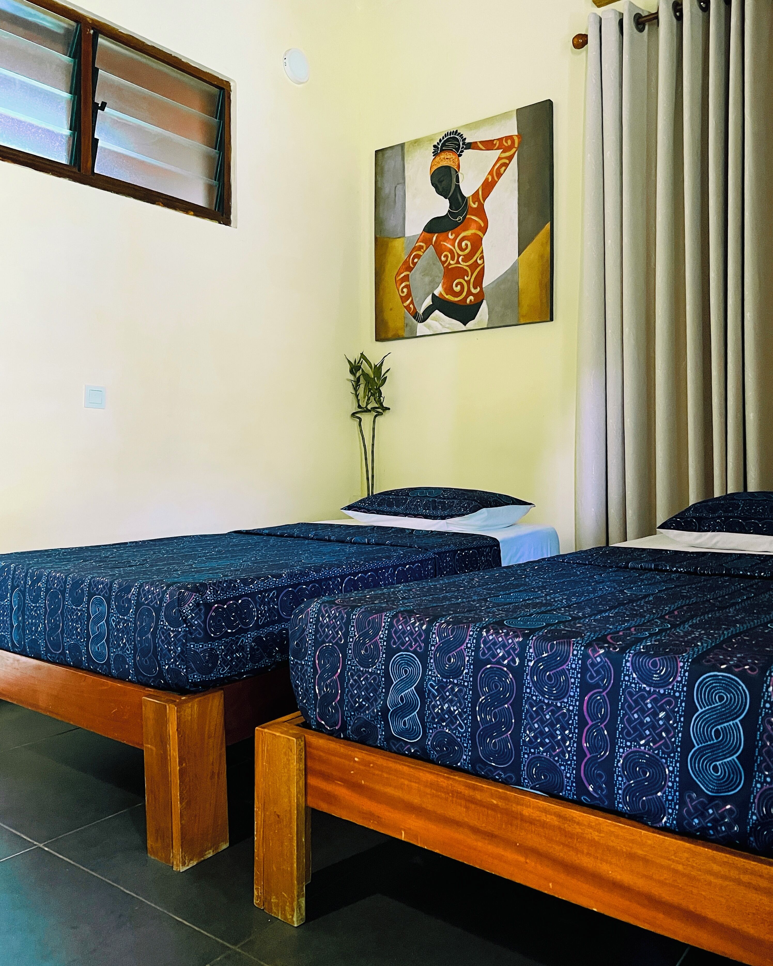 A room with two wooden beds with blue patterned bedding, a painting on the wall, a window with blinds, and a curtain.