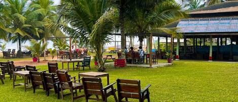 An outdoor seating area with wooden tables and chairs, surrounded by palm trees and a building with solar panels.