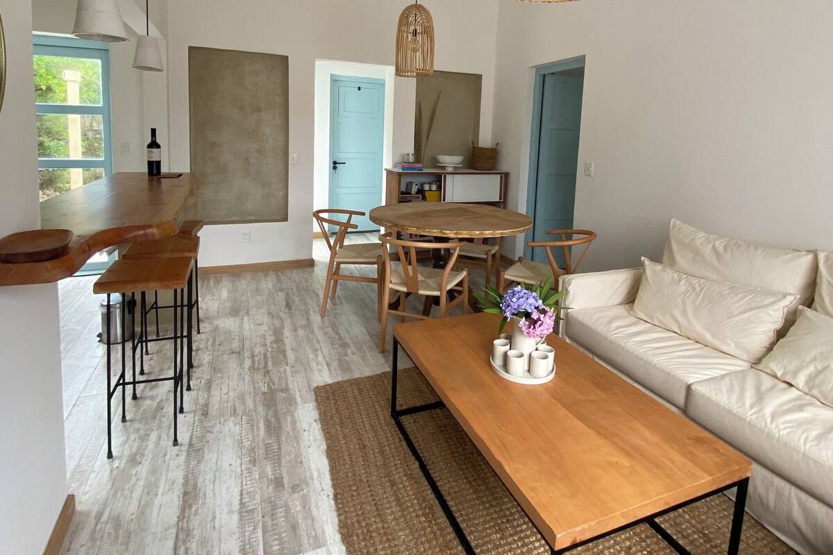 A spacious living area with a wooden coffee table, a sofa, and a dining table with chairs. There is a hanging wicker light fixture above the dining table.