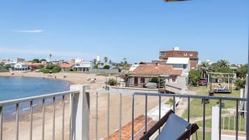 Uma varanda com duas poltronas de madeira, vista para a praia e um edifício com telhado vermelho ao fundo.