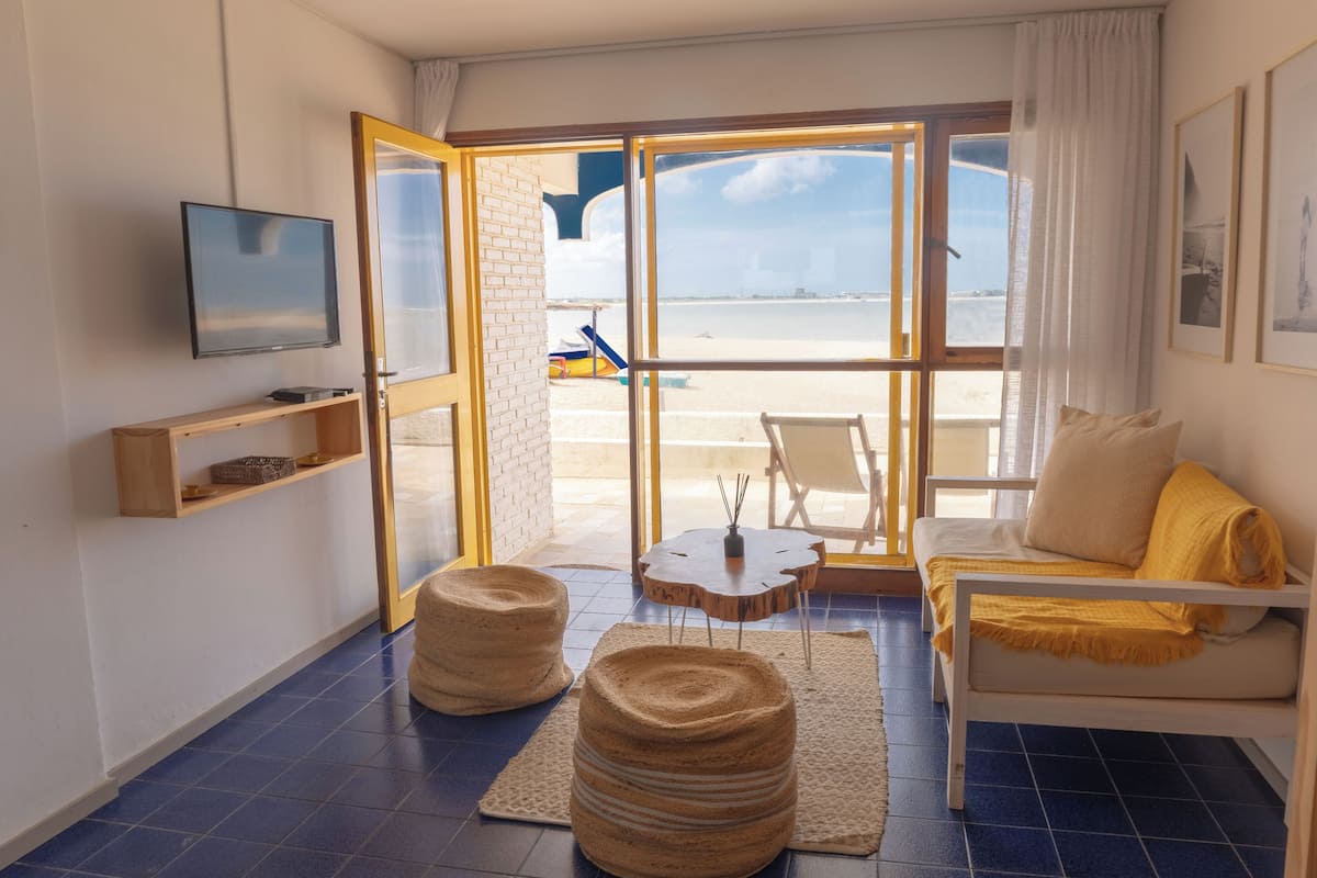A room with a large sliding glass door leading to a beach view, a white sofa with yellow cushions, a small round table, and a mounted TV.
