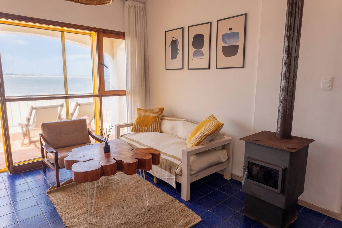 A cozy room with a sofa, a wooden coffee table, a fireplace, and a large window overlooking the sea.