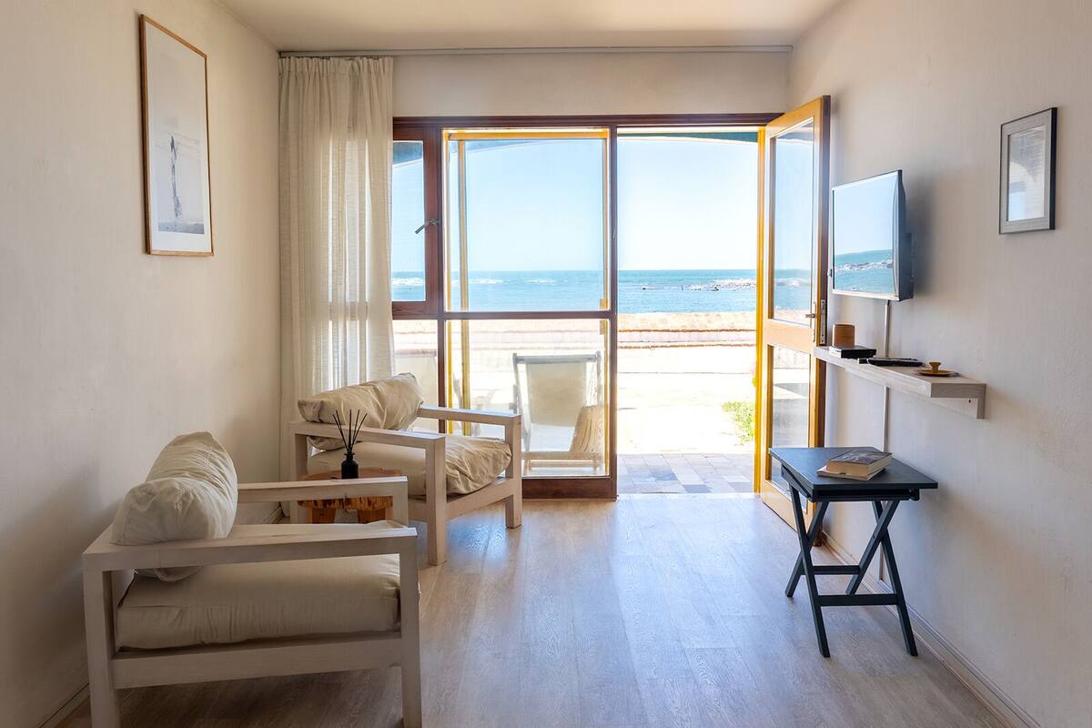 A room with a large window overlooking a beach, a sofa, a chair, a small table, and a TV on the wall.