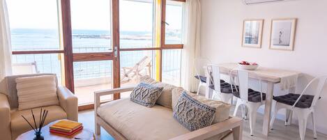 A spacious living area with a sofa, chairs, a dining table, and a view of the sea through large windows.
