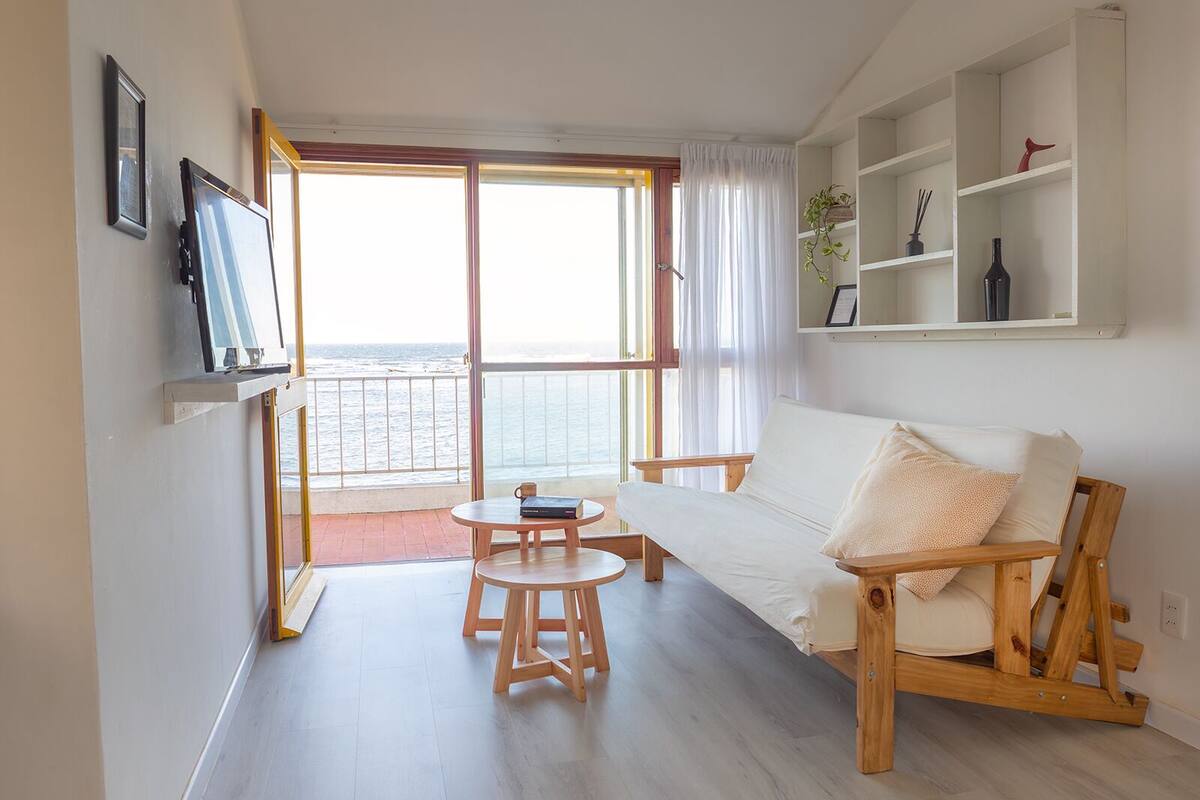 A living room with a wooden sofa, a small round table, and a TV mounted on the wall. There is a balcony with a view of the sea.