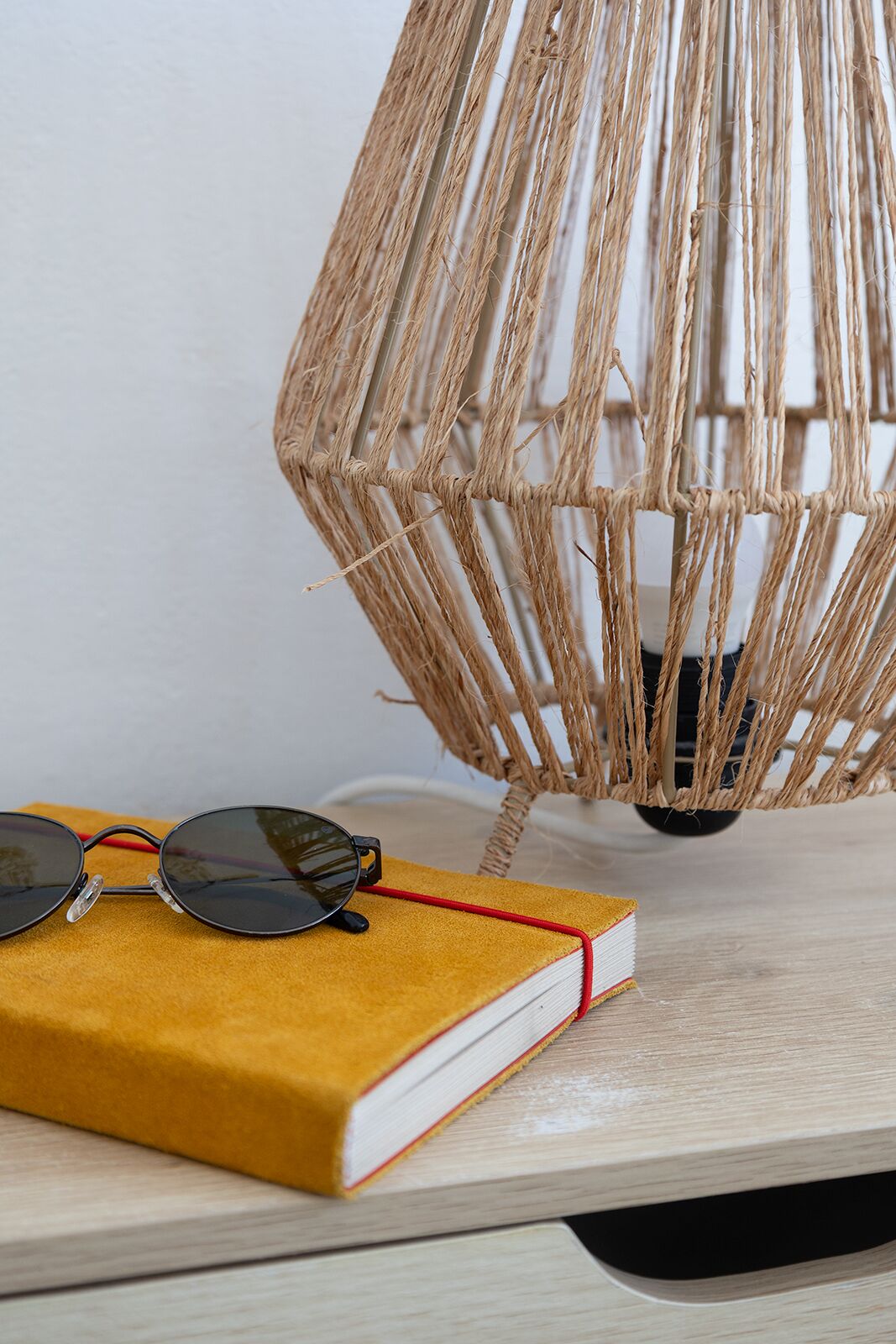 Una lámpara de techo trenzada, un par de gafas de sol y un libro amarillo sobre una superficie de madera.
