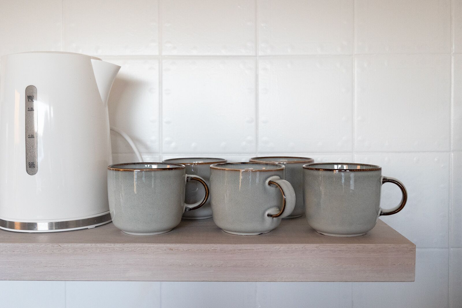 Una tetera blanca y cuatro tazas grises sobre un estante de madera frente a una pared de azulejos blancos.