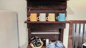 A kitchen shelf with a pink pitcher, colorful mugs, a coffee maker, and a plate of instant coffee packets.