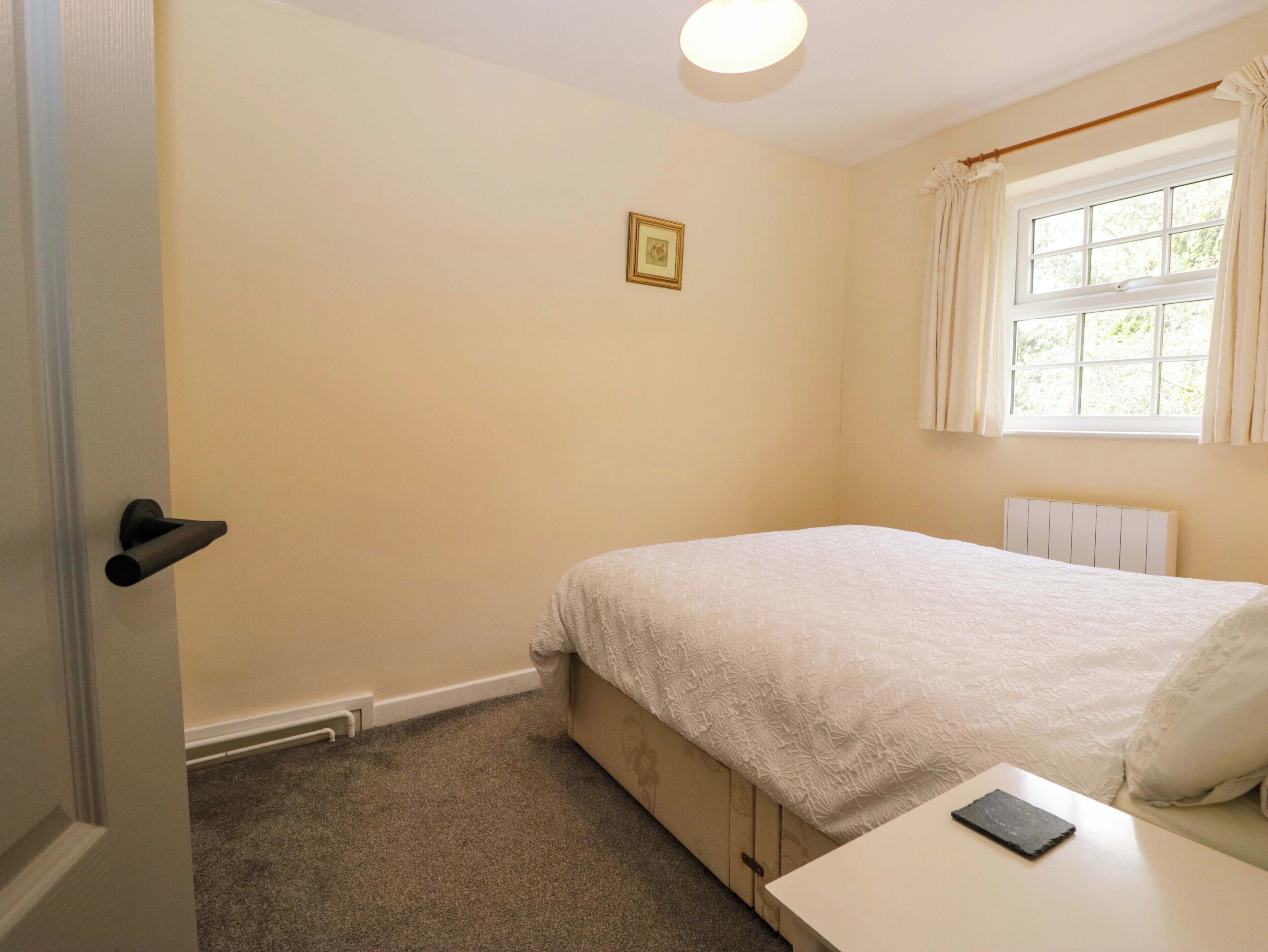 A bedroom with a single bed, a bedside table, a radiator, a window with curtains, and a door.