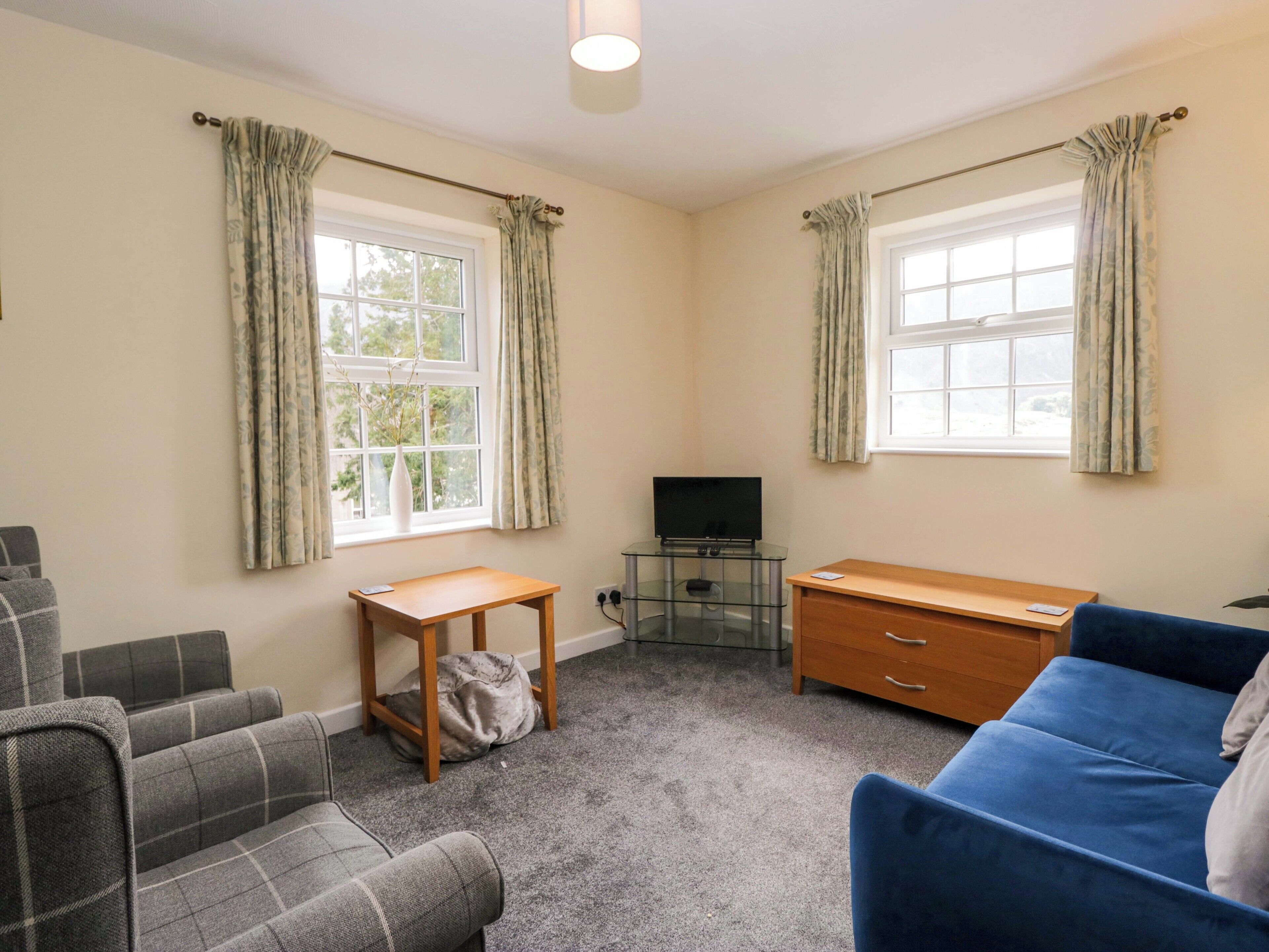 A room with two windows, a television on a glass table, a wooden desk, a blue sofa, and a grey sofa.