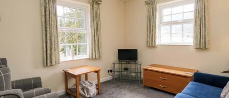 A room with two windows, a television on a glass table, a wooden desk, a blue sofa, and a grey sofa.