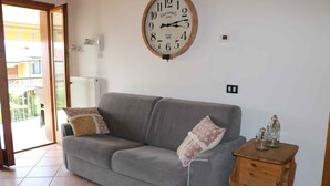 A living room with a grey sofa, two patterned throw pillows, a wooden side table with glass jars, and a wall-mounted vintage-style clock.