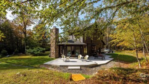 A modern house with a stone chimney, a covered patio with outdoor seating, and a well-maintained lawn. - Dreamwood: A Modern Catskills Retreat (Jeffersonville)